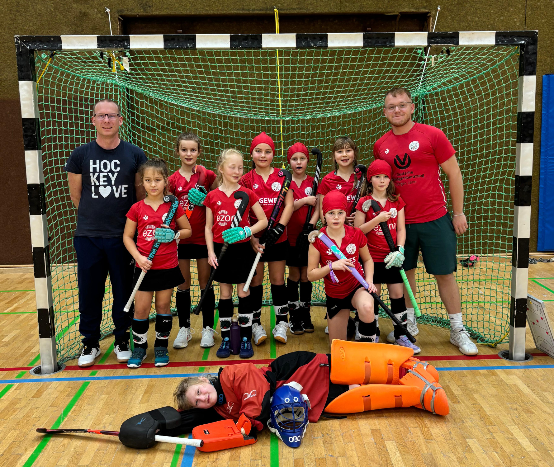 Hockeymannschaft in der Halle, mit Trainer und Kindern in roten Trikots.