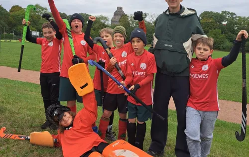Gruppe Kinder in rot-orangenen Trikots mit Coach auf Rasen, grauer Himmel.