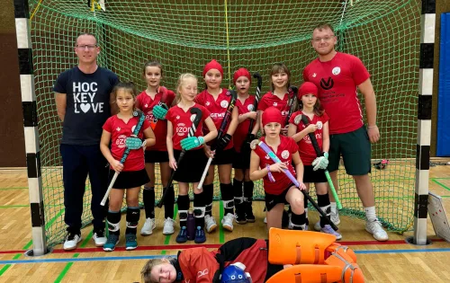 Hockeymannschaft in der Halle, mit Trainer und Kindern in roten Trikots.