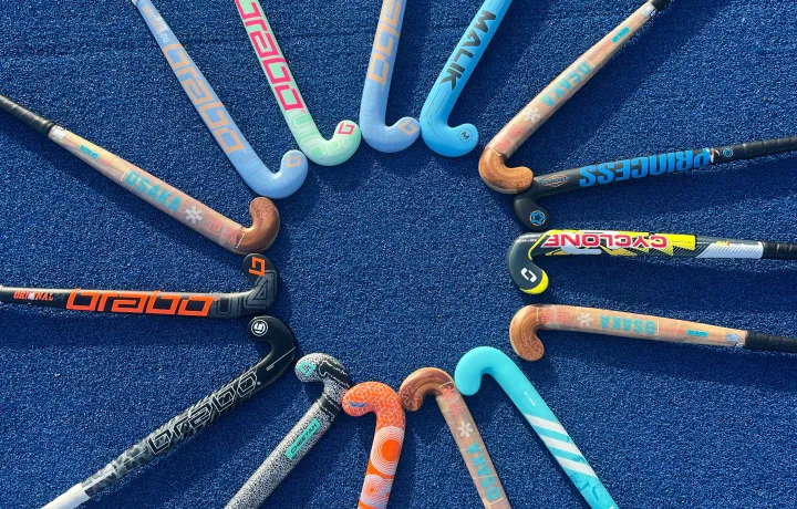 Mehrere bunte Hockeyschläger liegen fächerförmig im Kreis auf einem blauen Kunstrasenfeld, sodass die Schlägerköpfe in der Mitte zusammentreffen.