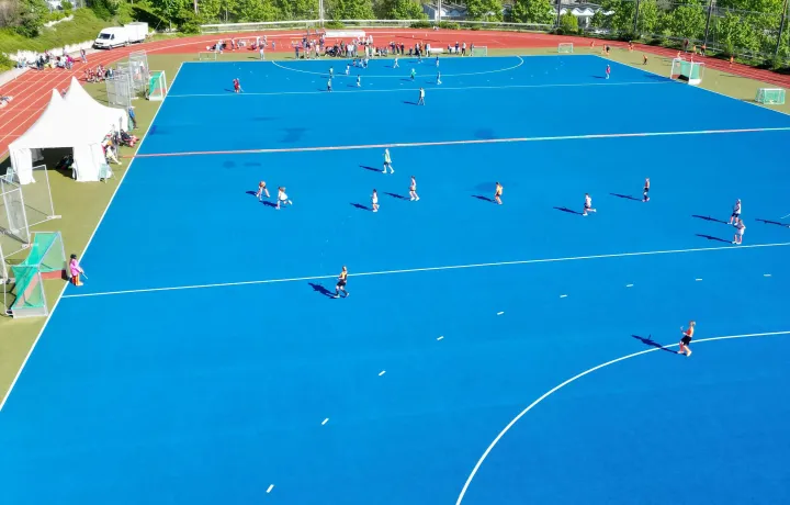 Luftaufnahme eines blauen Kunstrasen-Hockeyfeldes, auf dem mehrere Kinder in zwei Teams ein Spiel austragen. Am Spielfeldrand stehen Zuschauer, Zelte und Betreuer. Im Hintergrund sind Bäume, Gebäude und eine hügelige Landschaft unter klarem blauem Himmel zu sehen.