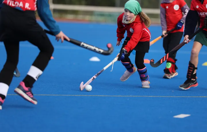 Junge Feldhockeyspielerin in rotem Trikot spielt den Ball auf blauem Kunstrasen, umgeben von weiteren Kindern mit Schlägern.