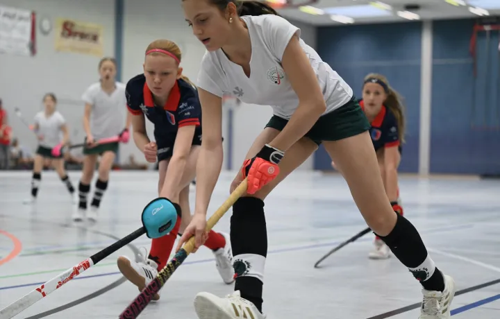 Hallenhockey-Szene: Spielerin in weißem Trikot spielt den Ball, eine Gegenspielerin in dunklem Trikot greift an; weitere Spielerinnen im Hintergrund.
