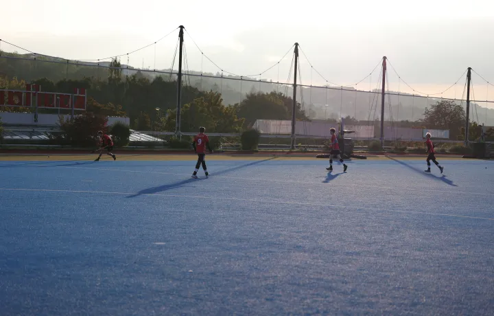 Mehrere Feldhockeyspieler in roten Trikots laufen über einen blauen Kunstrasenplatz; im Hintergrund Netze und Abendlicht.