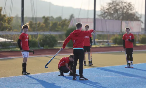 Jugendliche Feldhockeyspieler in roten Trikots stehen auf dem Platz; ein Trainer gibt Anweisungen, während ein Spieler am Boden sein Material richtet.