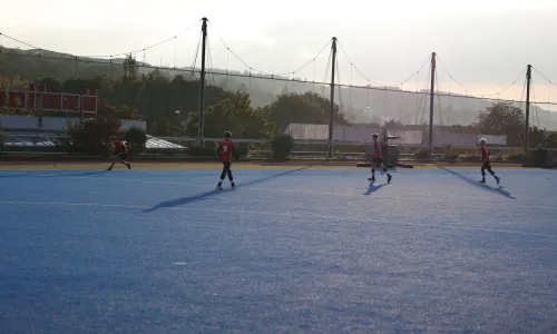 Mehrere Feldhockeyspieler in roten Trikots laufen über einen blauen Kunstrasenplatz; im Hintergrund Netze und Abendlicht.
