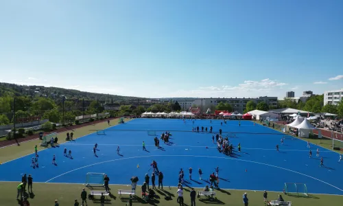 Weitwinkelaufnahme eines großen blauen Hockeyplatzes mit vielen Spielergruppen und Zuschauern; im Hintergrund Zelte und Stände bei sonnigem Wetter.
