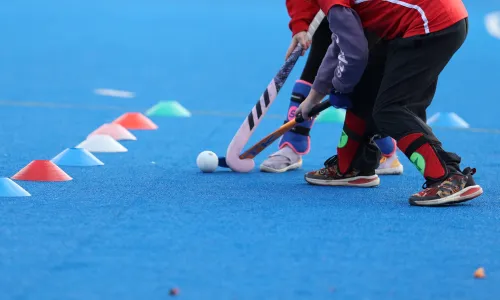 Zwei Kinder beim Hockeytraining auf blauem Kunstrasen, die mit ihren Schlägern einen Ball zwischen farbigen Markierungshütchen spielen.