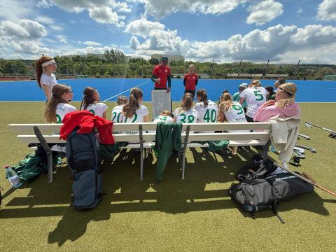 Eine Hockey-Mannschaft sitzt auf einer Bank am Spielfeldrand, während zwei Trainer vorne vor einem Taktikboard Anweisungen geben. Die Spielerinnen tragen weiße Trikots mit grünen Nummern, Hockeyschläger und Taschen liegen am Boden. Im Hintergrund ist ein blaues Kunstrasenfeld, Bäume und ein leicht bewölkter Himmel zu sehen.